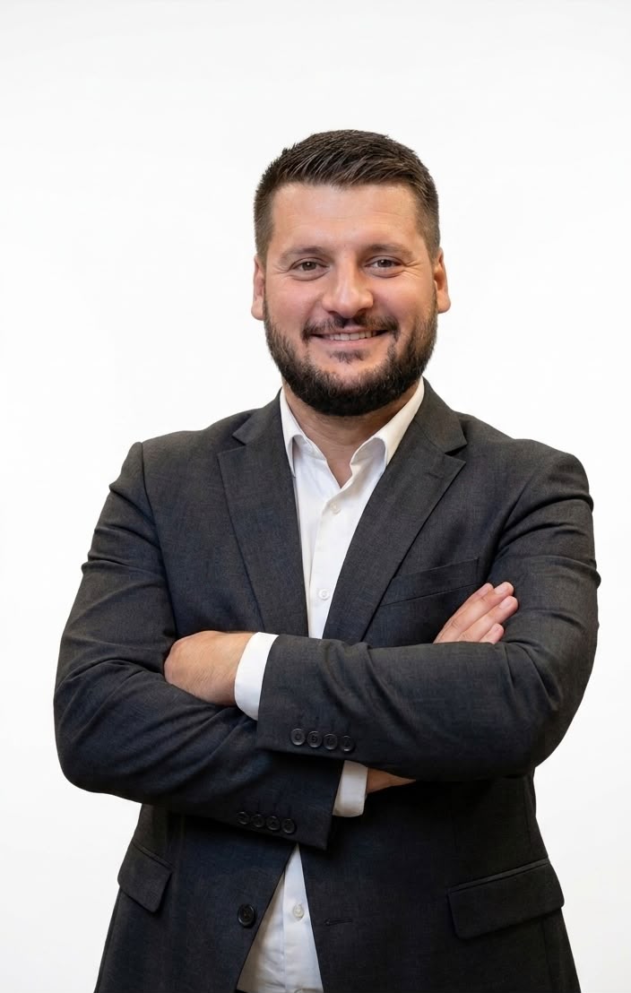 A professional portrait of Kushtrim Dragaj, a senior academic leader with a confident and encouraging expression. He is wearing a sharp charcoal suit in a minimalist modern office setting with soft, high-key lighting. The style is clean, corporate, and authoritative, reflecting the Level-Up Academy brand of excellence and stability.